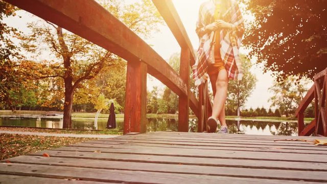 Woman Walking crossing Wooden Bridge in the Autumn Park. 4K. Unrecognizable young woman wearing denim sneakers shoes enjoying calm evening outdoors. Close up on boots. 