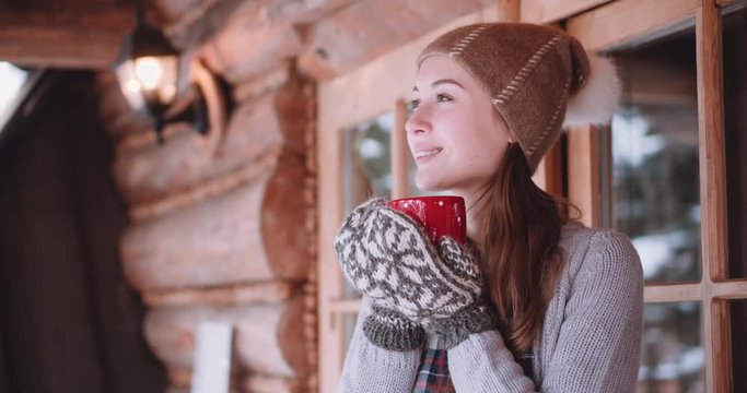 Woman with Cup of Hot Tea or Coffee Stands by the Cozy House on Snowy Winter Morning. 4K DCi SLOW MOTION 120 fps. Beautiful Girl Enjoying Winter Outdoors with a Mug of Warm Drink. Christmas Holidays 