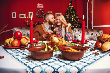 Beautiful couple kissing and holding glass of wine in a decorated festive interior with a Christmas tree. A romantic dinner for thanksgiving with fried chicken and candles