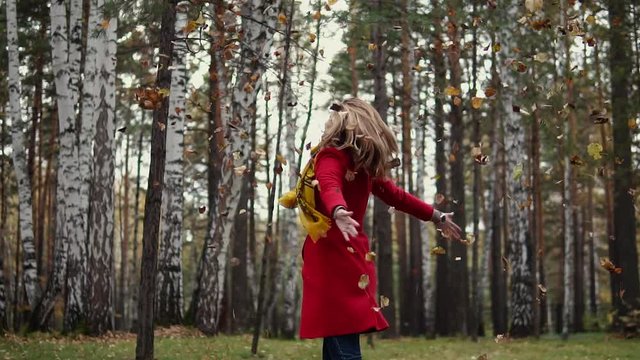 young woman throws up fallen yellow leaves and having fun. the girl in the red coat in the autumn forest. slow motion