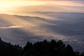 Obraz premium Aerial view of a valley, with sun rays coming out behind some mountains and hills at sunset, and trees in the foreground