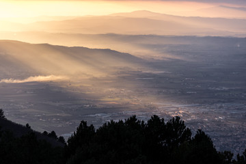 Aerial view of a valley, with sun rays coming out behind some mountains and hills at sunset, and trees in the foreground