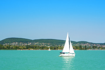 A view from a ship to bright Balaton lake water and a white yacht with a town, forest and mountains at the background on sunny summer day, Hungary.