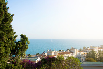 A view to Mediterrain sea from a view point over the tile roofs of white houses and hotels, green trees and pink violet colorful bougainvillea flowers at the foreground.