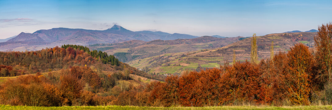 gorgeous autumn countryside panorama in mountains. forest with red foliage on rolling hills, village in valley and mountain ridge with high peak in a distance. Beautiful landscape in Carpathians