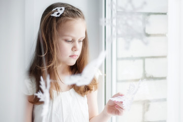 Beautiful girl near the window holds a paper snowflake in her hands