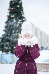 A girl stands on the street in front of a Christmas tree and blows on the snow in her arms