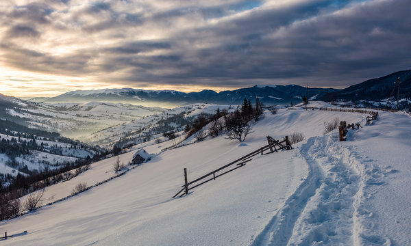 beautiful countryside in mountains. path in a snow near the wooden fence over the village and rural fields on hillsides covered with snow at sunrise