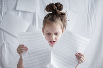 A little girl sits among blank sheets of paper and looks at the torn sheet of paper in her hands