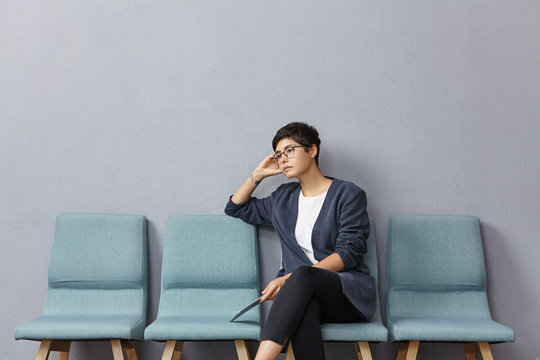 Sad Upset Female Job Seeker Being Thick And Tired Of Finding Work, Sits On Chair Before Talk With Emloyer, Has Thoughtful Expression, Holds Modern Tablet Computer, Thinks What To Tell About Herself