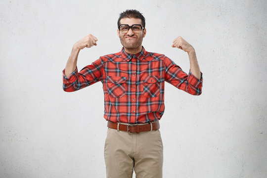 Funny ridiculous bearded guy wearing eyeglasses with thick lenses and checkered red shirt posing in studio raising arms with pumped fists, tensing muscles to show his big biceps and strength
