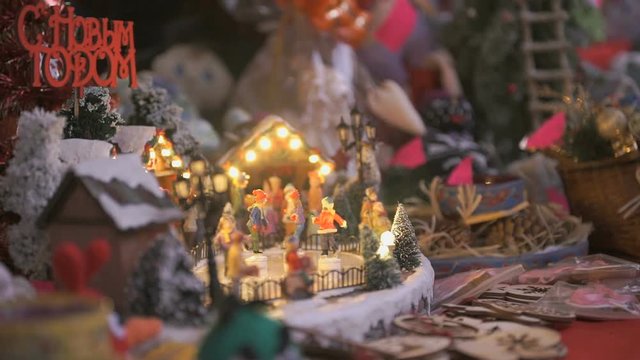 Close-up - miniature souvenir Christmas toy - little people skate in circle on an ice rink illuminated with garland with winter scenery with the inscription in Russian happy NEW YEAR.