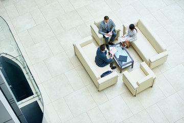 Directly above view of concentrated managers in formalwear analyzing financial figures while having informal working meeting at modern spacious office lobby