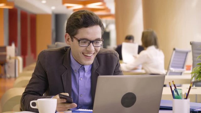 young happy man gets great news about success and triumph during busy working day with papers sitting at table in co working colorful business office space