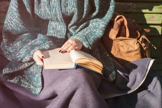 Woman in autumn knitted clothes (poncho) covered with warm plaid reading outside, sitting on wooden seesaw