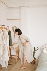 Attractive brunette young woman with short hair in linen beige robe at home standing in her white room in morning in front of rail with clothes and chooses outfit for day.