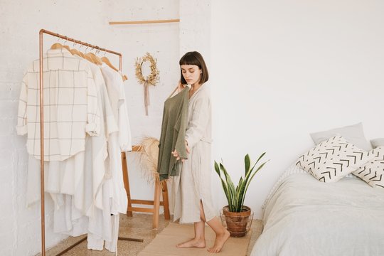 Attractive brunette young woman with short hair in linen beige robe at home standing in her white room in morning in front of rail with clothes and chooses green sweater to wear.
