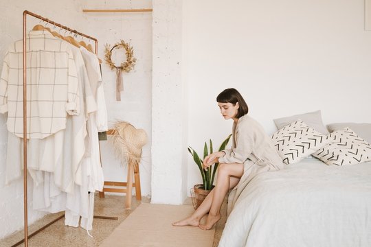 Young sexy lady with short black hair in linen beige robe sits on bed in morning in her big white room, looks down and thinks what to wear today. Space for your text.