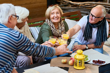 Group of smiling senior friends toasting with glasses of orange juice while celebrating momentous event at cozy outdoor cafe