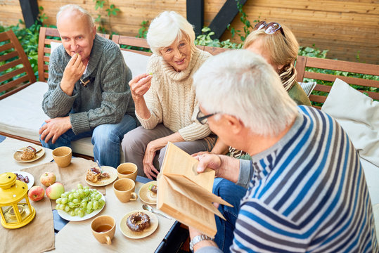 Group of joyful senior friends having fun outdoors: they enjoying delicious pastry and fragrant coffee while elderly man in striped sweater reading their favorite novel aloud