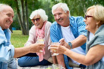 Two joyful senior couples toasting with glasses of orange juice while celebrating momentous event at green park