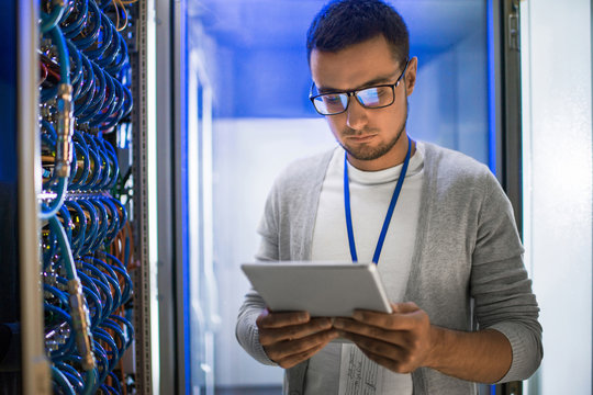 Portrait of young man using digital tablet standing by server cabinet while working with supercomputer in blue light