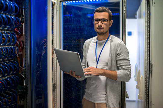 Portrait of young man holding laptop and looking at camera standing by server cabinet while working with supercomputer in blue light