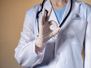 A female doctor wearing rubber gloves shows a gesture "OK". Female doctor with a stethoscope gesticulating on a beige background.