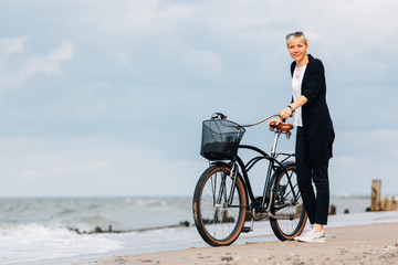 Young stylish blonde woman in black cardigan with pretty retro bicycle with basket on the beach. Handsome girl resting near ocean after ride
