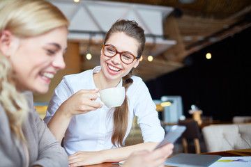 Cheerful girl with cup of tea laughing during talk to her friend in cafe