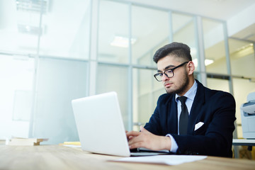 Young broker or manager in suit networking in office in front of laptop