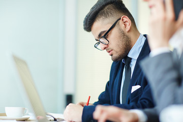 Busy young man in formalwear making notes with colleague near by