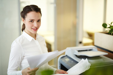 Young secretary with papers standing by photocopier in office
