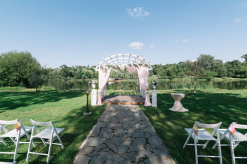 Beautiful summer wedding ceremony outdoors. Decorated chairs stand on the grass. Wedding arch made of light cloth and white and pink flowers on a green natural background. Wooden doors. Empty seats