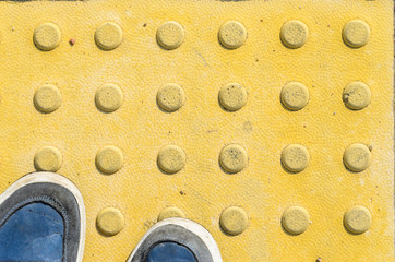 View from the First Person to his Feet, Standing in Front of the Sidewalk with Tactile Tiles to Navigate Blind People. Blue Sneakers on Yellow Tactile Paving Slabs.