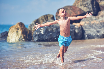 Cute Caucasian boy is running in the water along the sea shore. His arms are wide open.