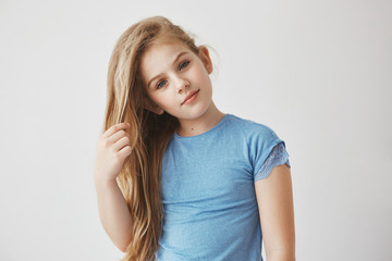 Obraz premium Close up portrait of beautiful little blonde girl with blue eyes holding hair with hand, looking in camera with relaxed and calm face expression.