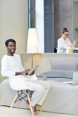 Peaceful weekend at home: handsome young man posing for photography with toothy smile while browsing Internet on digital tablet and listening to music in headphones, his girlfriend seen on background