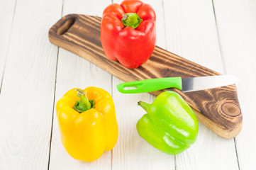 Row of red yellow and green fresh whole raw pepper on rural cutting board beside acute knife on old white rustic planks