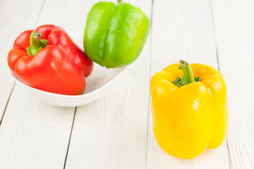 Single yellow fresh whole raw pepper beside green and red pepper in ceramic bowl on old white rustic planks