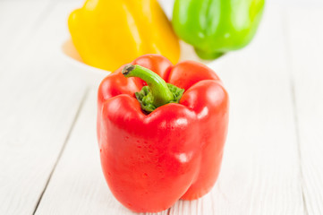 Single red fresh whole raw pepper beside green and yellow pepper in ceramic bowl on old white rustic planks