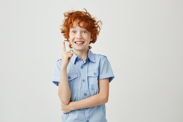 Portrait of cheerful little boy with ginger hair and freckles smiling, pointing upside with finger,...