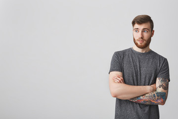 Beautiful young bearded caucasian man with dark hair and tattooed arm crossing hands, looking aside...