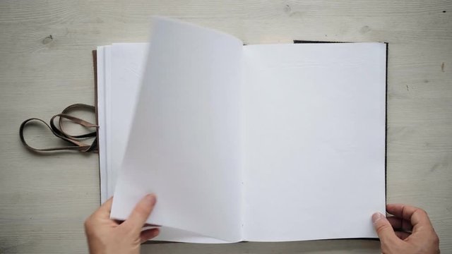 Man's hands turning over clean blank pages from recycled paper in big notebook, isolated on white wooden table , then closing it, wrapping around with leather cord and taking away