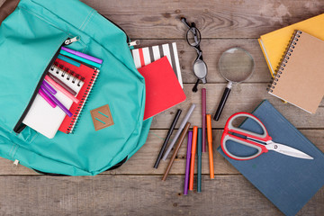 Backpack and school supplies: books, magnifying glass, notepad, felt-tip pens, eyeglasses, scissors on brown wooden table