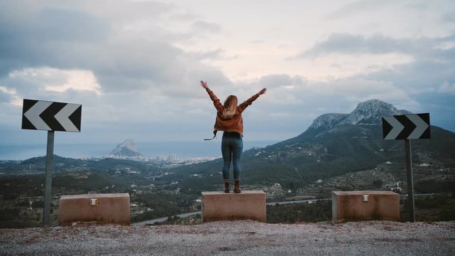 Camera gets closer to woman that stands on side of road on top of hill or summit, raising arms into air as sign or concept of business success or personal win, excited and proud