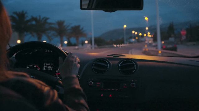 Interior view of a woman driving her car through the outskirts of city or residential neighborhood in twilight or sunset hours, traffic lights passing by, hands on steering wheel