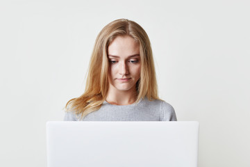 Indoor shot of serious concentrated young blonde female with appealing appearance, reads information attentively online or watches video, uses laptop computer. Technology and lifestyle concept