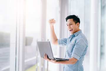 Happy excited Asian man holding laptop and raising his arm up to celebrate success or achievement.
