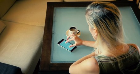 Dolly a blonde drinks cofee and reading an article in the phone while sitting at a table in the kitchen.